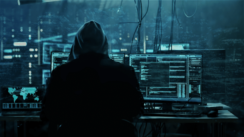 image of a hacker in a hoodie sitting infront of multiple computers